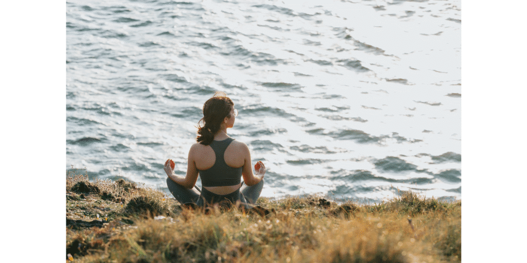 A woman sitting in a meditative yoga pose on a grassy bank, looking out over rippling water. This scene represents the importance of stress management and regular movement—such as walking along Port Perry’s waterfront trails—for overall mental and physical health.