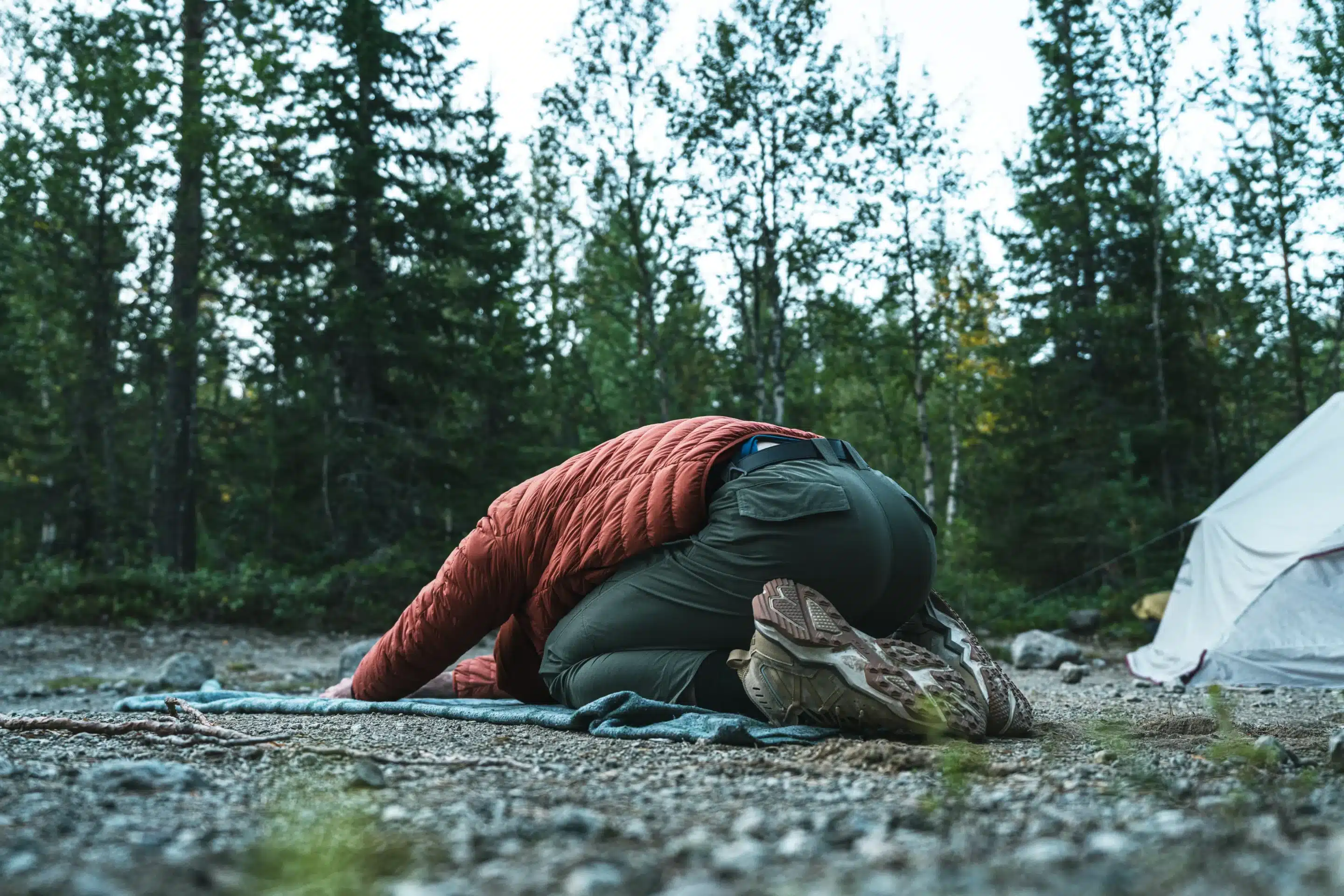 A person wearing a quilted orange jacket and green hiking pants performs a child's pose stretch on a blue mat outdoors. They are at a campsite with a white tent nearby and a dense forest in the background.