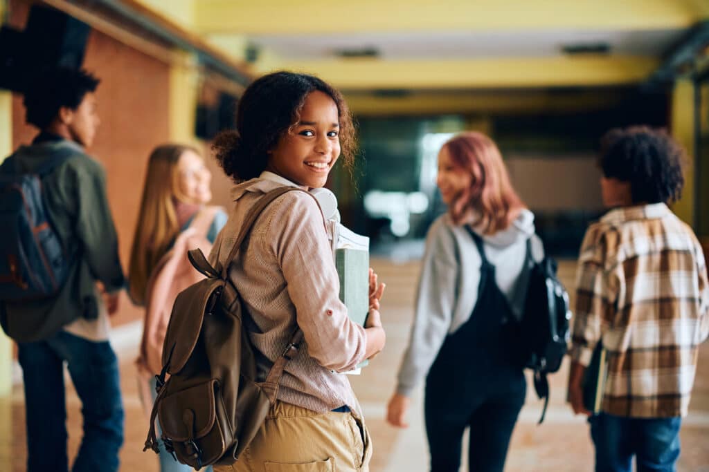 Happy African American high school student walking through hallway with her friends and looking at camera.