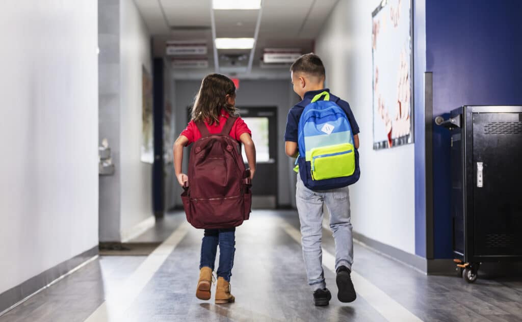 Rear view of two multiracial elementary students walking in a school hallway carrying backpacks, conversing. The girl is mixed Asian and Hispanic. The boy is Hispanic. They are 6 years old.