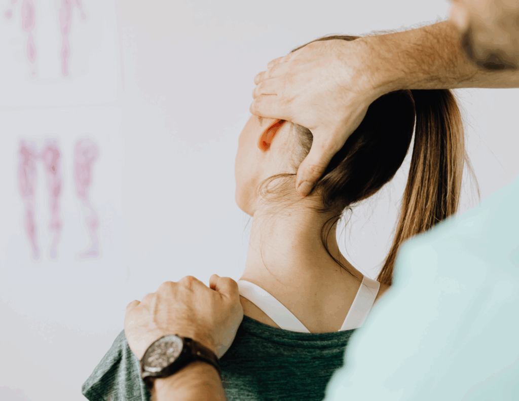 A close-up, back view of a woman sitting while a practitioner performs a cervical assessment. The practitioner has one hand on her shoulder and the other supporting the base of her head and neck. In the background, a blurred anatomical chart is visible on the wall.