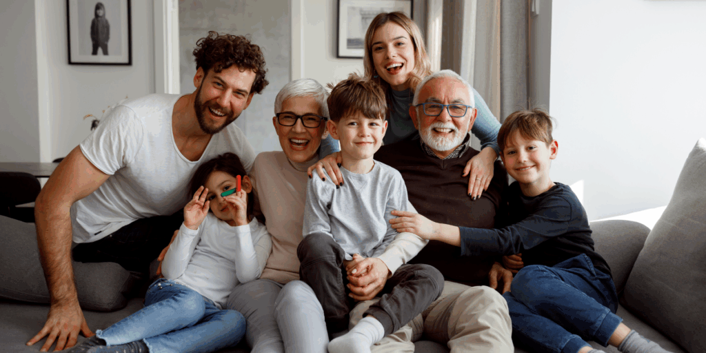 A happy multi-generational family of seven sitting together on a gray couch. The group includes grandparents, parents, and three young children, all smiling warmly at the camera in a bright living room.