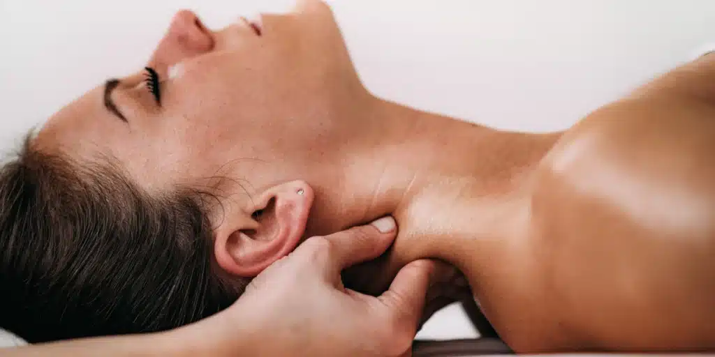 A close-up, profile view of a person lying down during a therapeutic treatment. A practitioner’s hand is gently applying pressure to the side of the person’s neck, just below the ear and jawline.