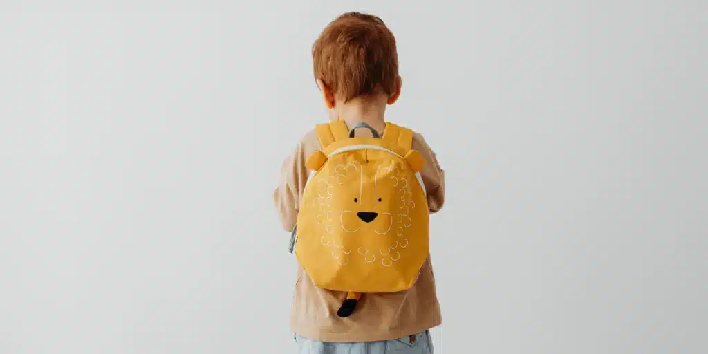 Rear view of a young child with reddish-brown hair standing against a light gray wall. The child is wearing a tan t-shirt and a bright yellow backpack shaped like a lion’s face, complete with small ears and a tail hanging from the bottom.