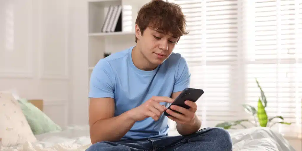 A teenage boy with wavy brown hair sits on a bed with a white comforter, focused on using a dark-colored smartphone. He is wearing a light blue t-shirt and jeans. The background shows a bright room with white window shutters and a bookshelf.