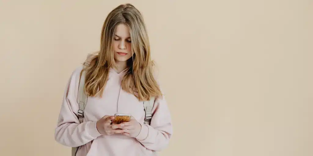 A teenage girl with long blonde hair stands against a plain beige background, looking down at a smartphone in her hands. She is wearing a light pink oversized hoodie and a beige backpack.