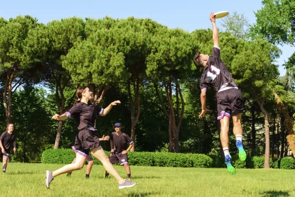 Action shot of a co-ed ultimate frisbee game on a grassy field surrounded by lush green trees. In the foreground, a male player wearing a black and purple jersey jumps high into the air with his right arm fully extended to catch a white flying disc, while teammate looks on from the grass.