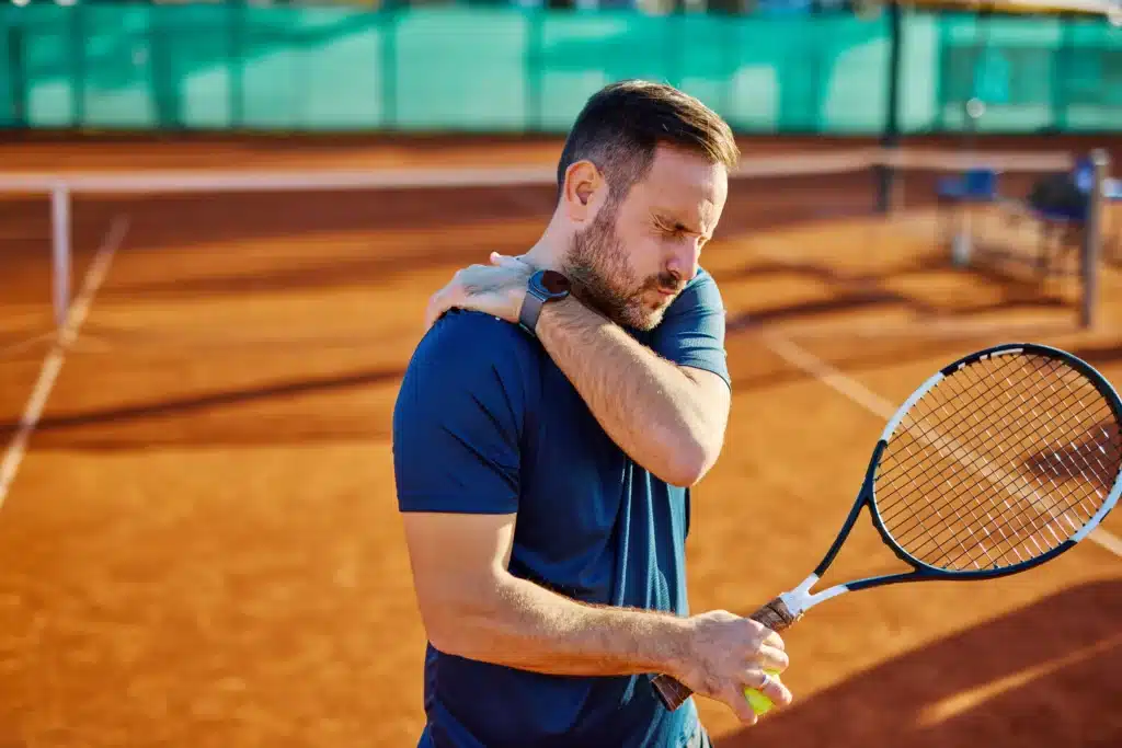 A male tennis player standing on an outdoor clay court, wincing in pain while gripping his right shoulder with his left hand. He is wearing a blue athletic shirt and holding a tennis racket and ball in his right hand, looking visibly uncomfortable from an apparent injury that requires chiropractic care.