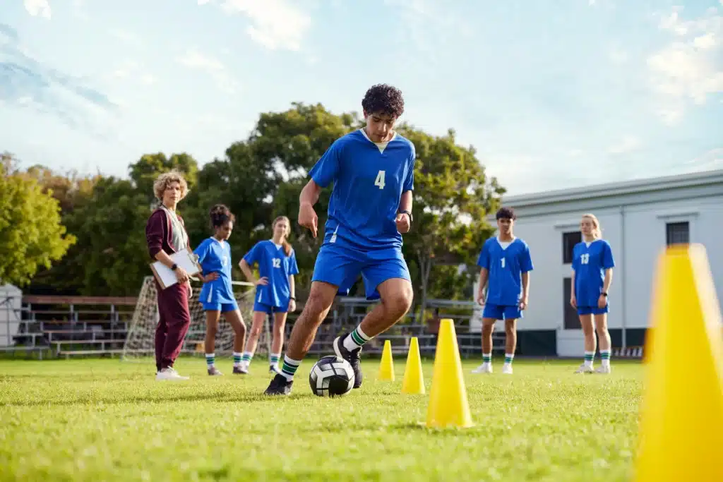 A young male soccer player in a blue uniform with the number 4 weaves a soccer ball through a line of bright yellow cones during a practice drill on a green field. A female coach holding a clipboard and several other teammates in matching blue jerseys stand in the background watching his progress.
