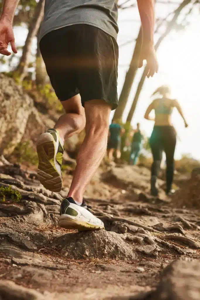 A low-angle, close-up shot of a trail runner’s legs and feet navigating a steep, rocky path with tree roots, while other runners follow behind in a sun-drenched forest.