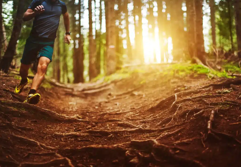A blurred, action-oriented shot of a male runner sprinting through a dense forest trail, with golden sunlight filtering through the tall trees in the background.